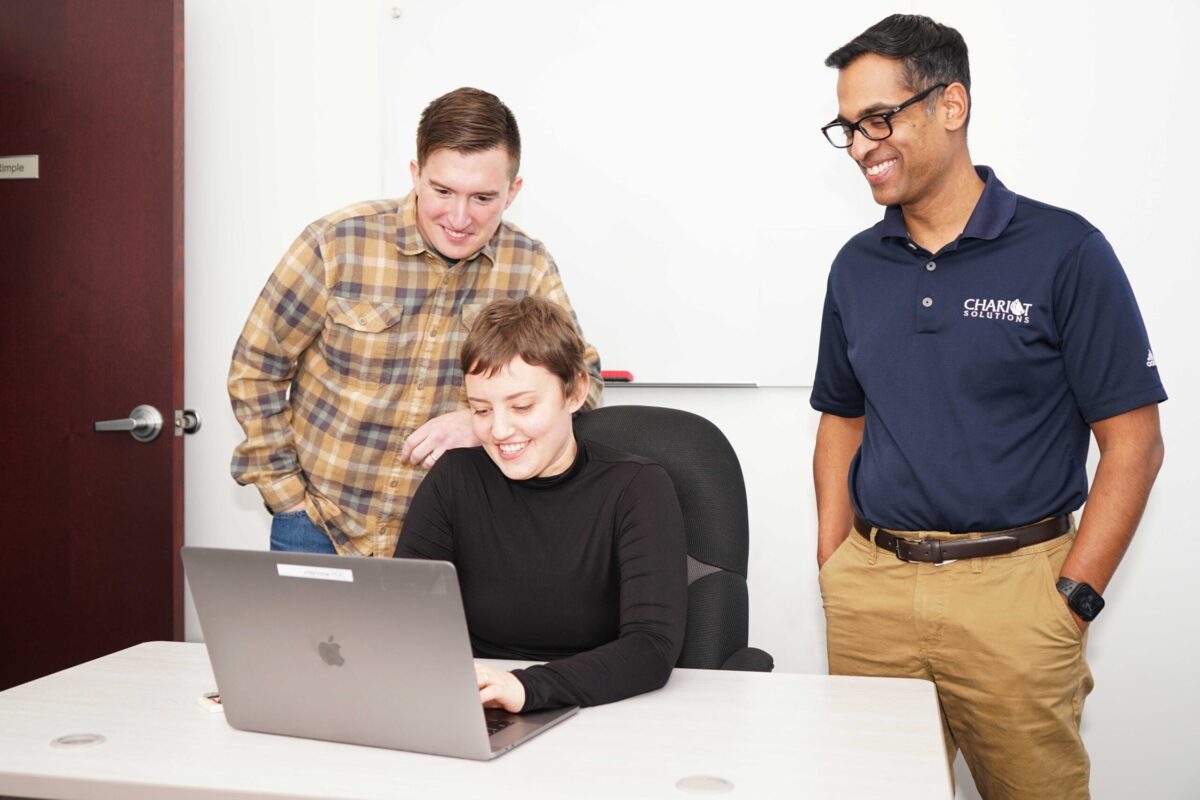 Photo of a woman sitting at a laptop while two men stand behind her smiling and helping.
