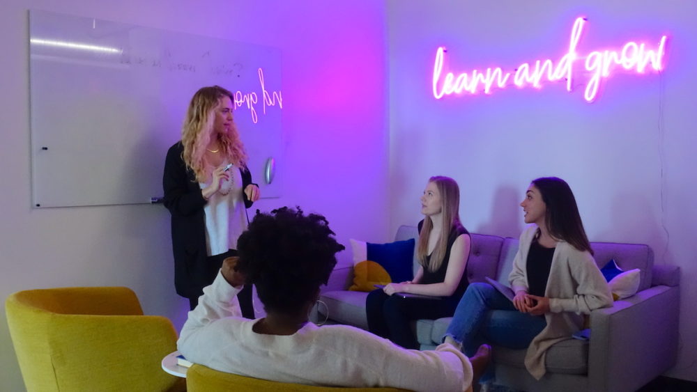 A group of women technologists in front of a neon sign that says "Learn and Grow"