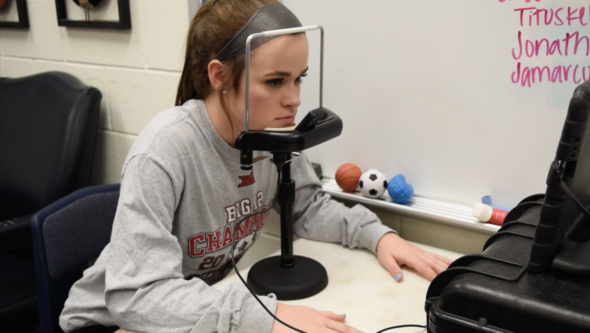A seated woman in a athletic clothing faces forward, her chin placed in a medical device that will track her eye movements.