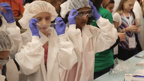 Two girls in lab coats and sterile gloves put on goggles.