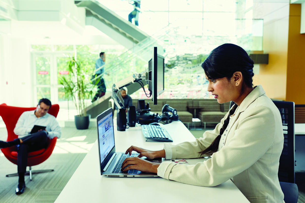 Photo of a woman working on a laptop in the foreground while other technologists work and walk past in the background.