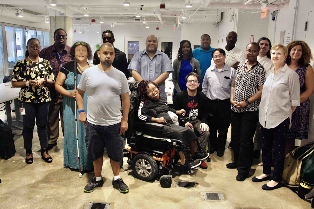 2GI participants standing or seated in wheelchair wearing multicolored clothing on brown floor.