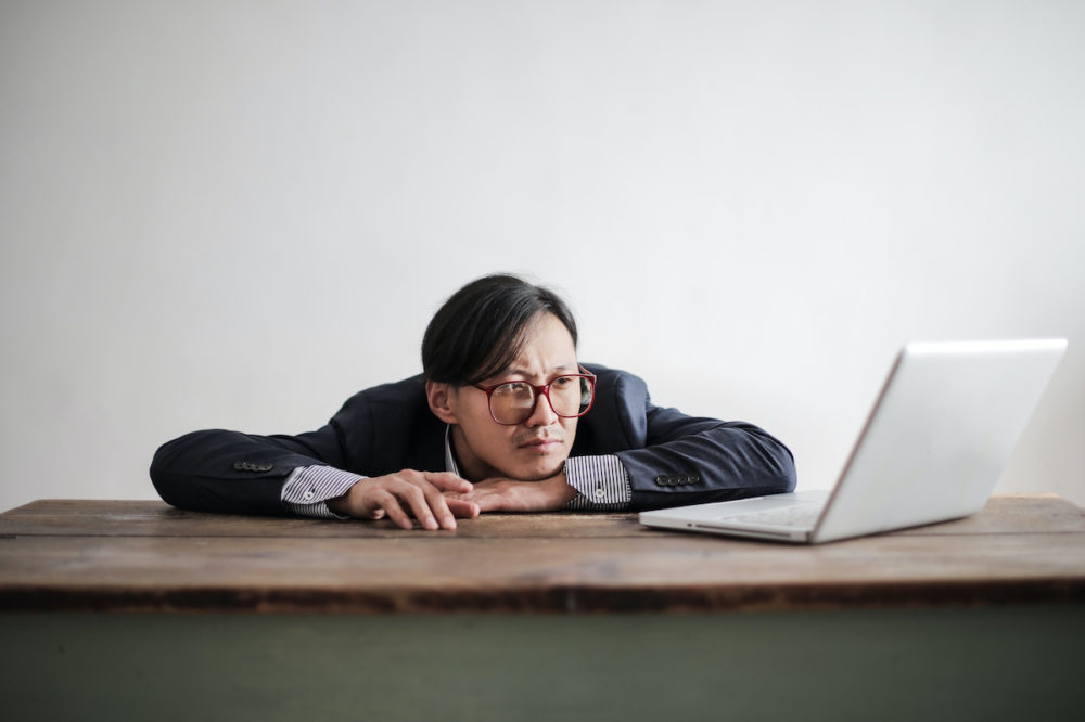 Man in grey suit with glasses places head on brown desk.