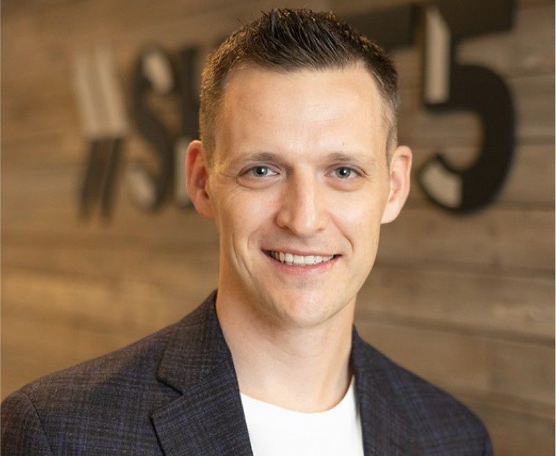 Man in charcoal suit and white t-shirt in front of brown wall reading "SHIFT5."