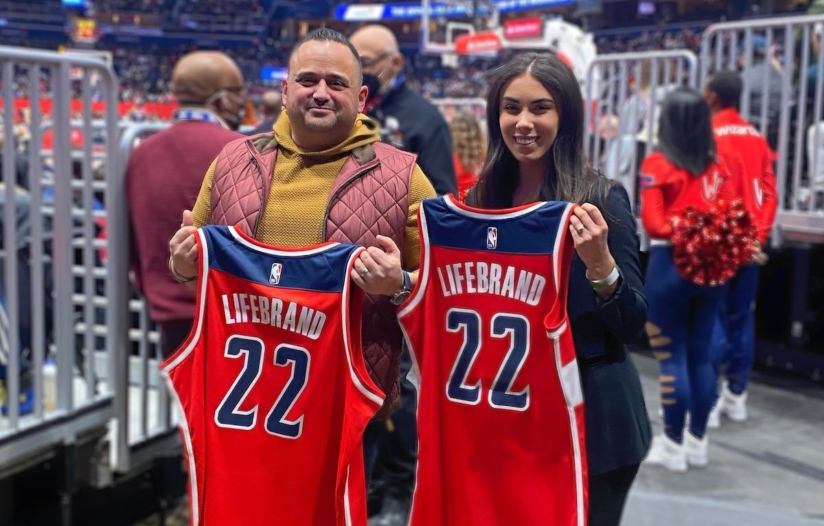 Two people stand in an arena, each holding a red basketball jersey with the name "Lifebrand" and the number "22" on the back. The crowd and court are visible in the background.