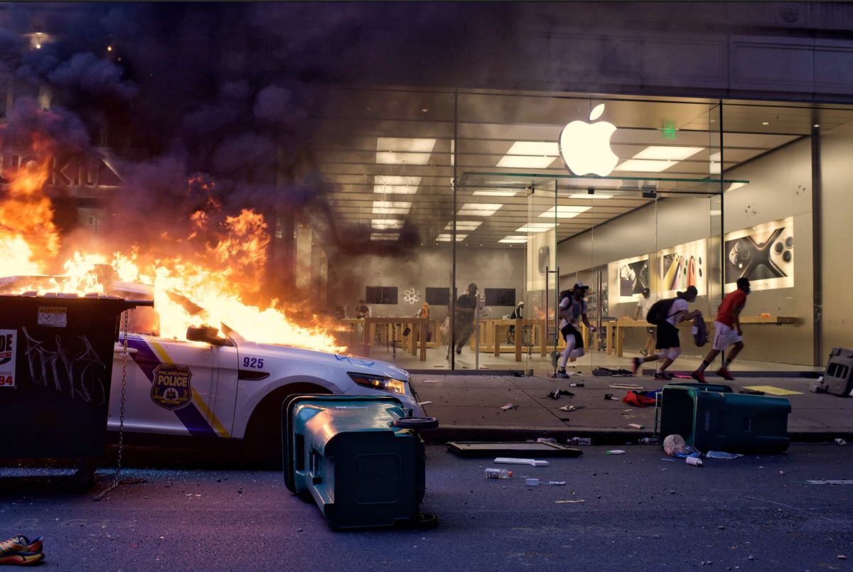 A police car is on fire outside an Apple Store as people run past, with smoke filling the street and debris scattered on the ground.