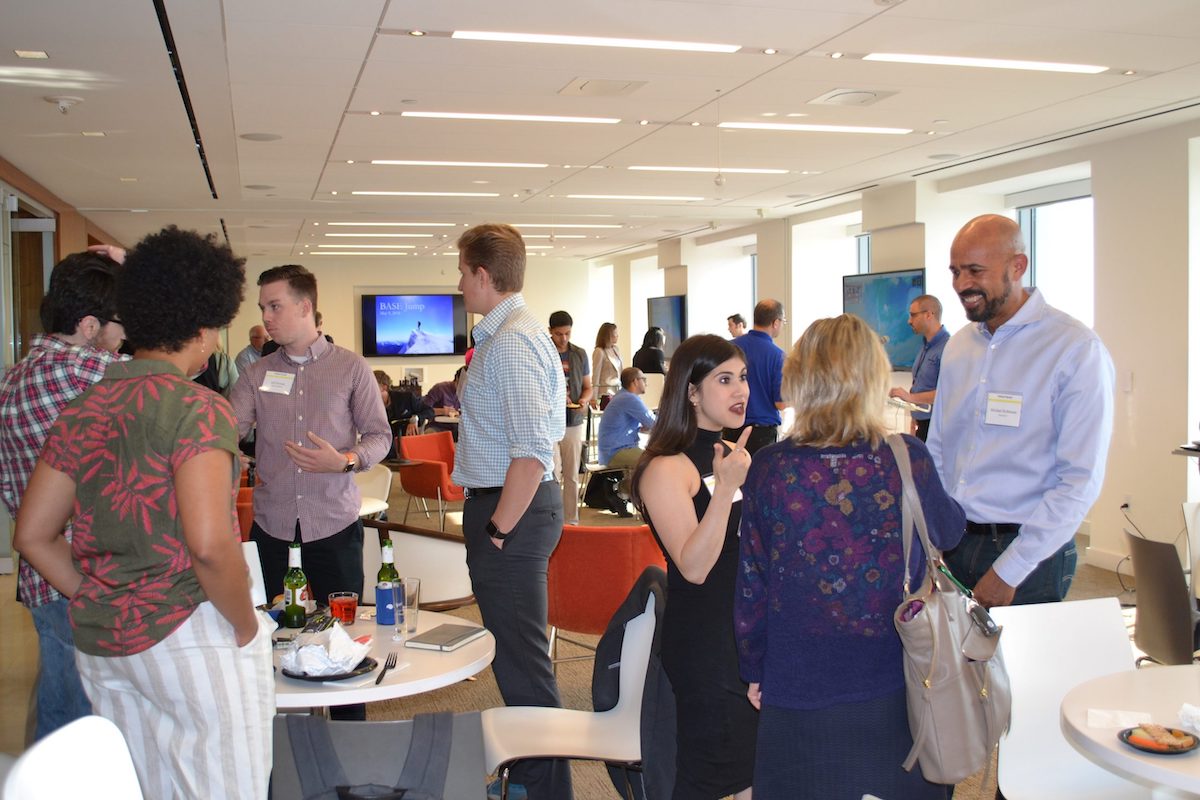 A diverse group of people engaging in conversation at an indoor networking event with tables, chairs, and screens in the background.