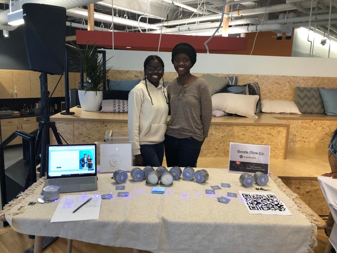 Two teen girls stand behind a table showcasing the candles they sell.