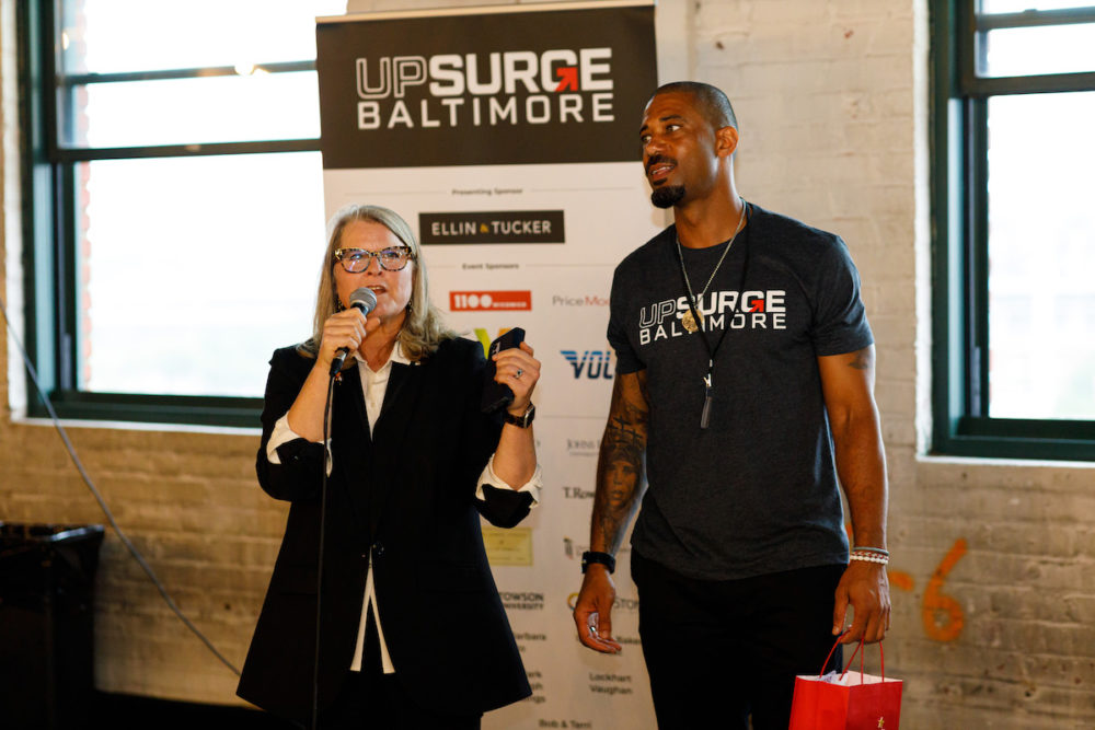 Woman in black suit with white shirt speaks next to man in black shirt and pants with white text near black, white and red sign and brown wall.