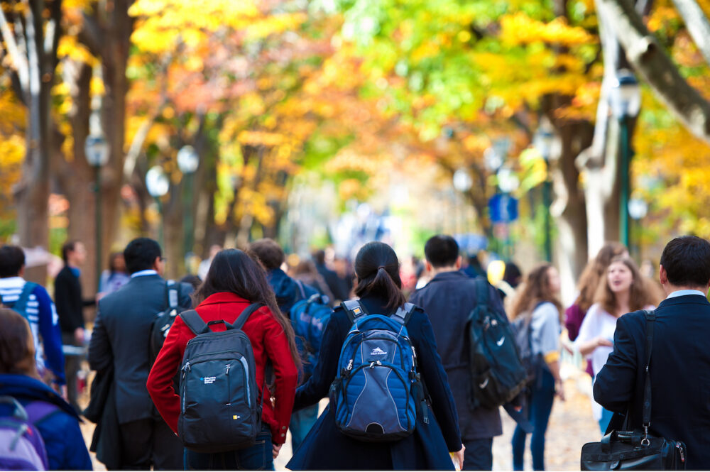 Crowd of students on UPenn's Locust Walk in multicolored clothing below brown trees with green and orange leaves.