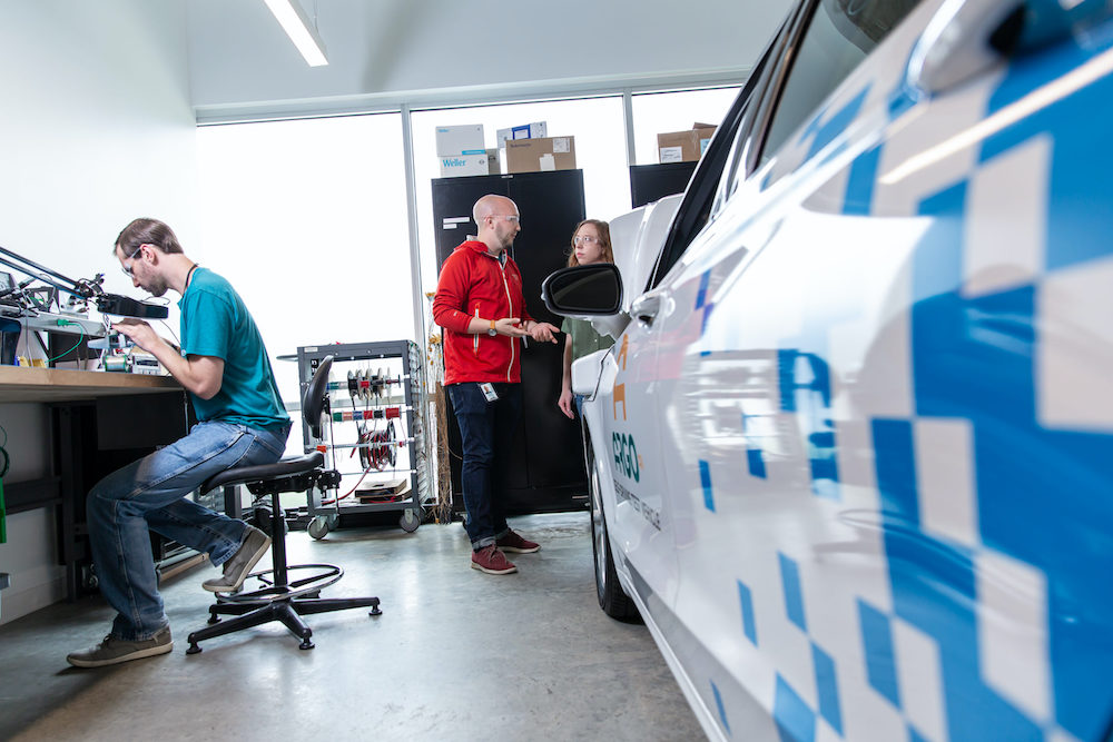 Technologists stand and talk near a white autonomous vehicle