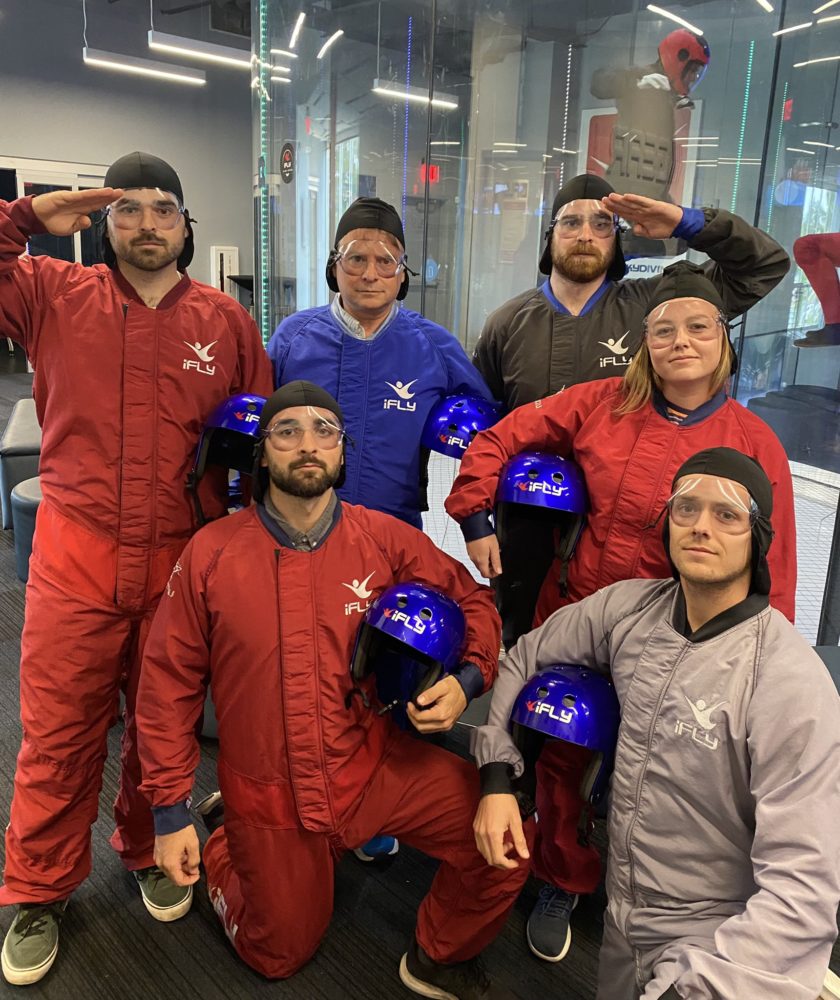 A group dressed in indoor skydiving suits and holding helmets poses at PAX Investor day.