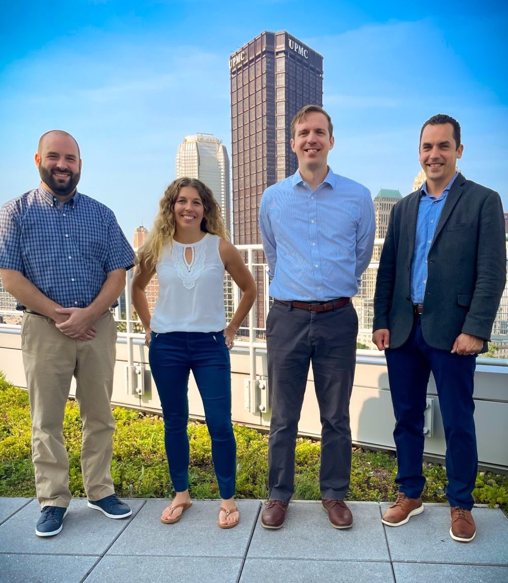 Four people stand side by side on a rooftop with city buildings, including UPMC and PPG towers, visible in the background on a clear day.