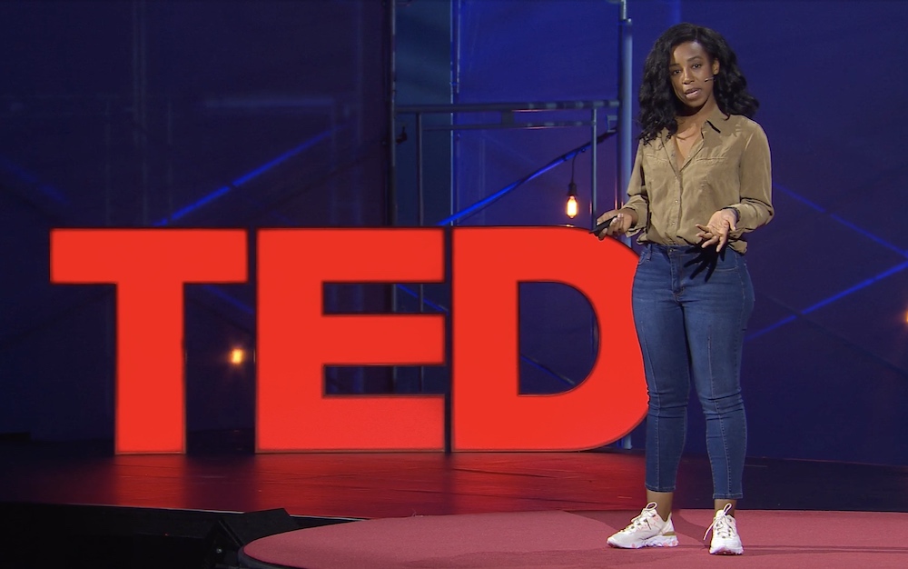 Woman in green shirt and blue jeans stand before red TED sign and blue wall