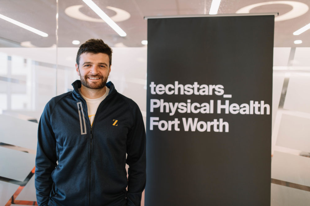 Brendan Sullivan stands before a sign that reads: techstars Physical Health Fort Worth
