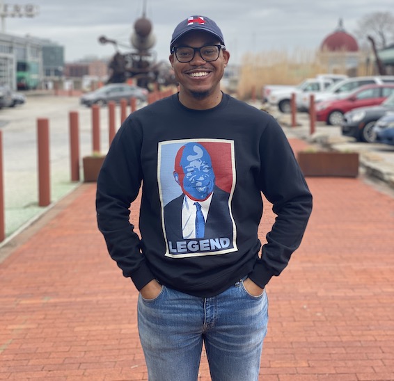 Man in navy hoodie with white and red images and blue jeans smiles on red walkway