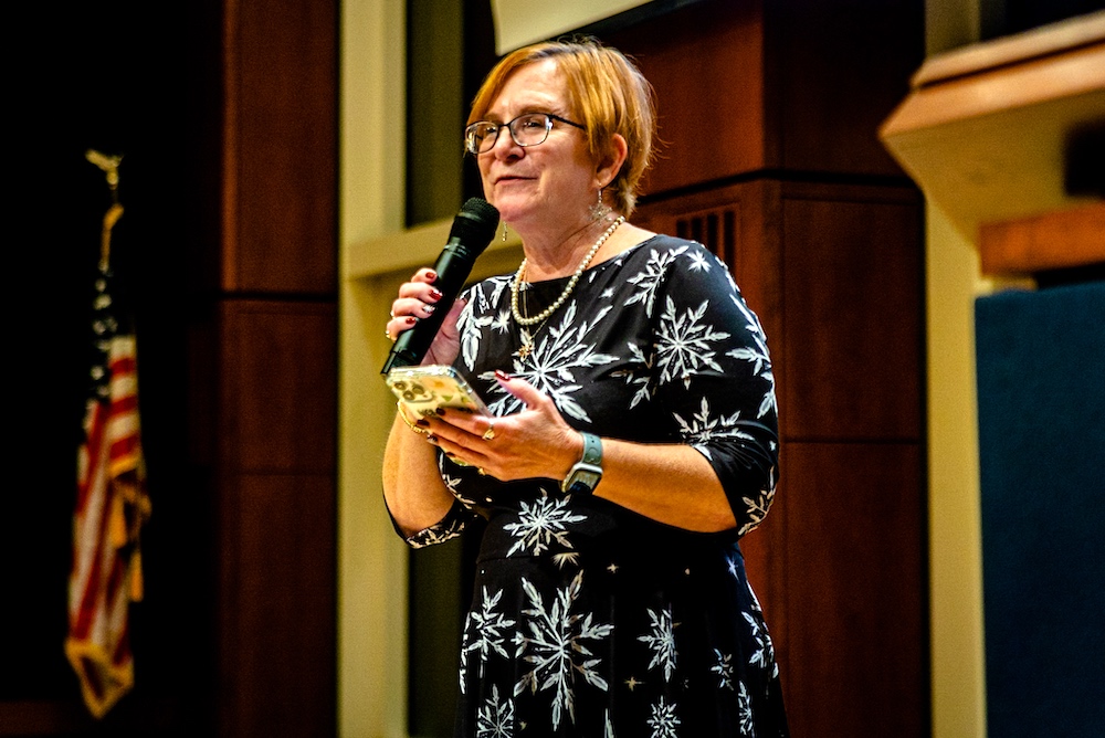 A woman with short hair and glasses holds a microphone and a phone while speaking on stage. An American flag is visible in the background.