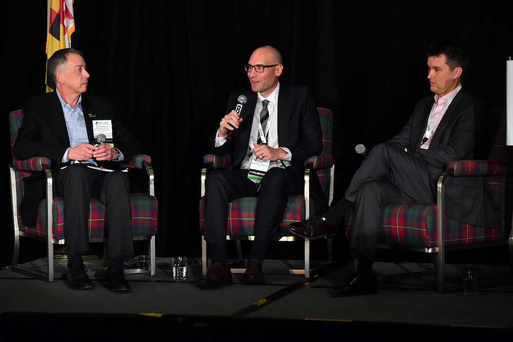 Three men in suits sit onstage and speak into microphones.