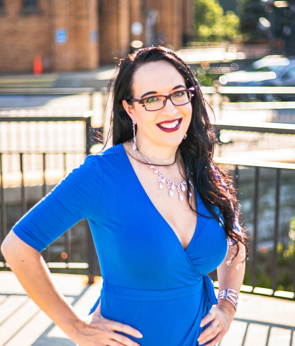 Woman with long dark hair and glasses wearing a blue dress and necklace, smiling with hands on hips, stands in an outdoor area with a brick building in the background.