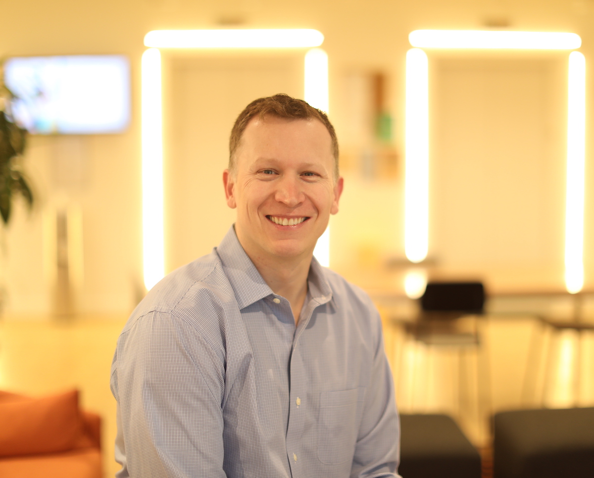 A man wearing a light blue shirt smiles at the camera in an indoor setting with blurred background lighting.