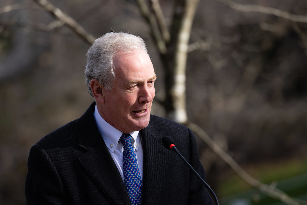 Man in black suit with blue tie speaks before beige trees.