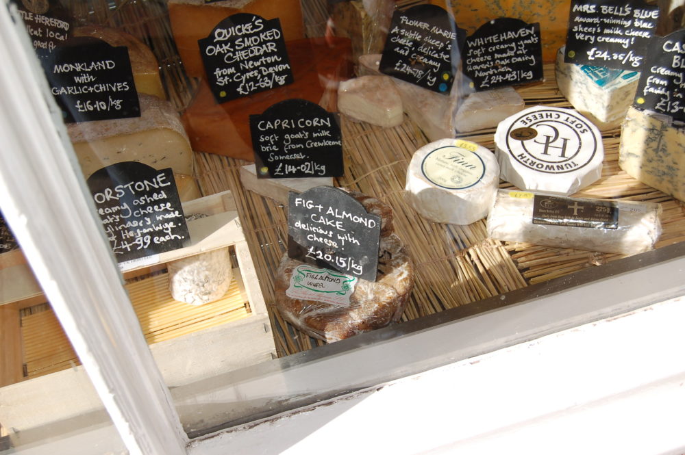 A cheese display on a wooden board featuring small cheese wheels and chalkboard signs with name and description.