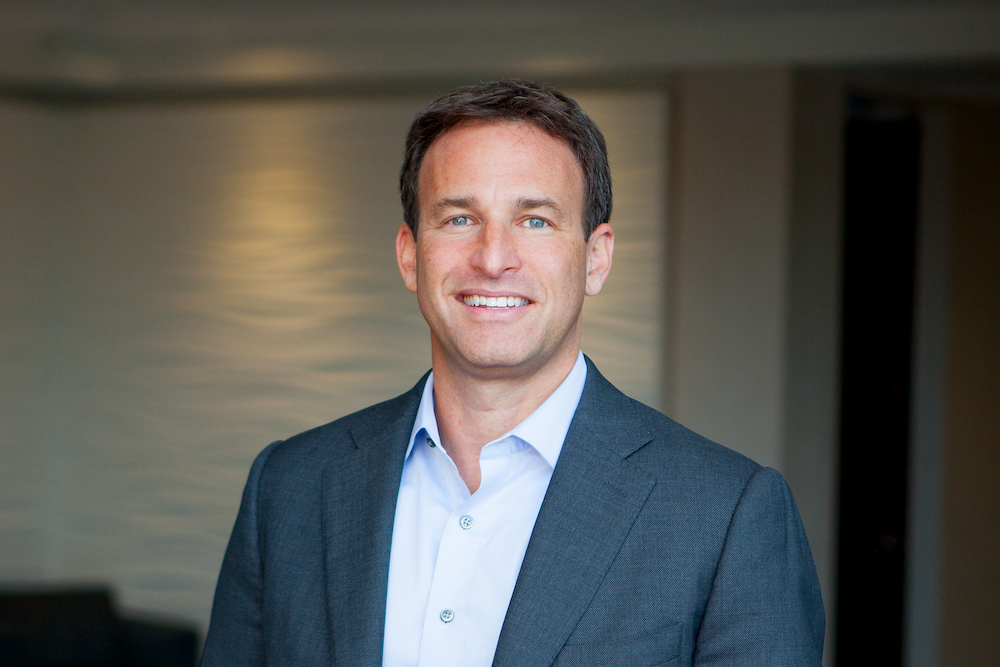 Chad Gottesman smiles in navy suit with light bule shirt in front of brown wall