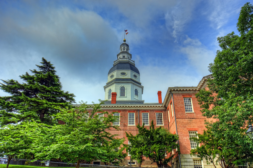 Maryland State House with white and green dome over red brick building by green trees and blue sky with white and grey clouds.