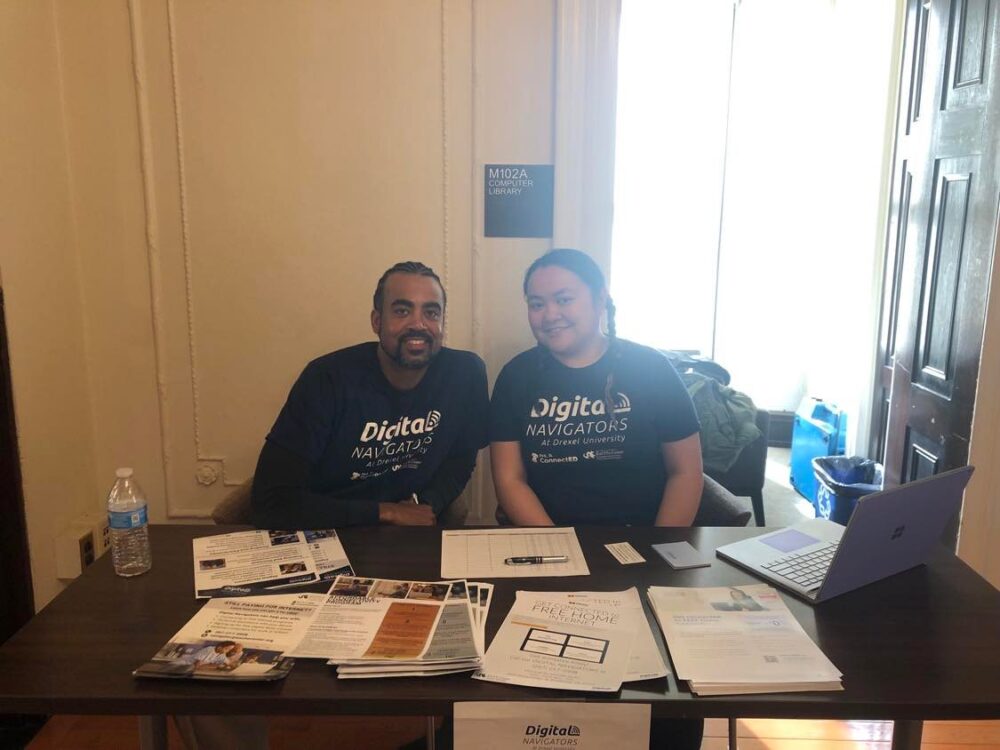 Two people wearing "Digital Navigators" shirts sit at an information booth with pamphlets, forms, a laptop, and a water bottle. A sign on the wall reads "M120A Information Commons.