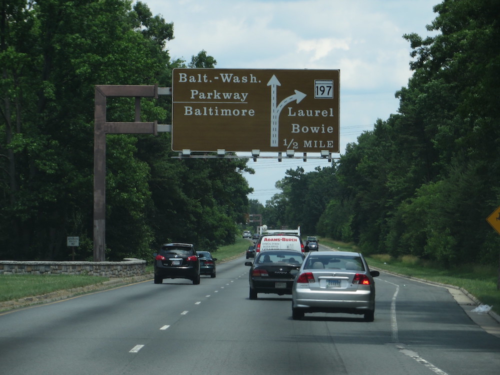 Brown highway sign with white text over cars and grey road with white lines near green trees