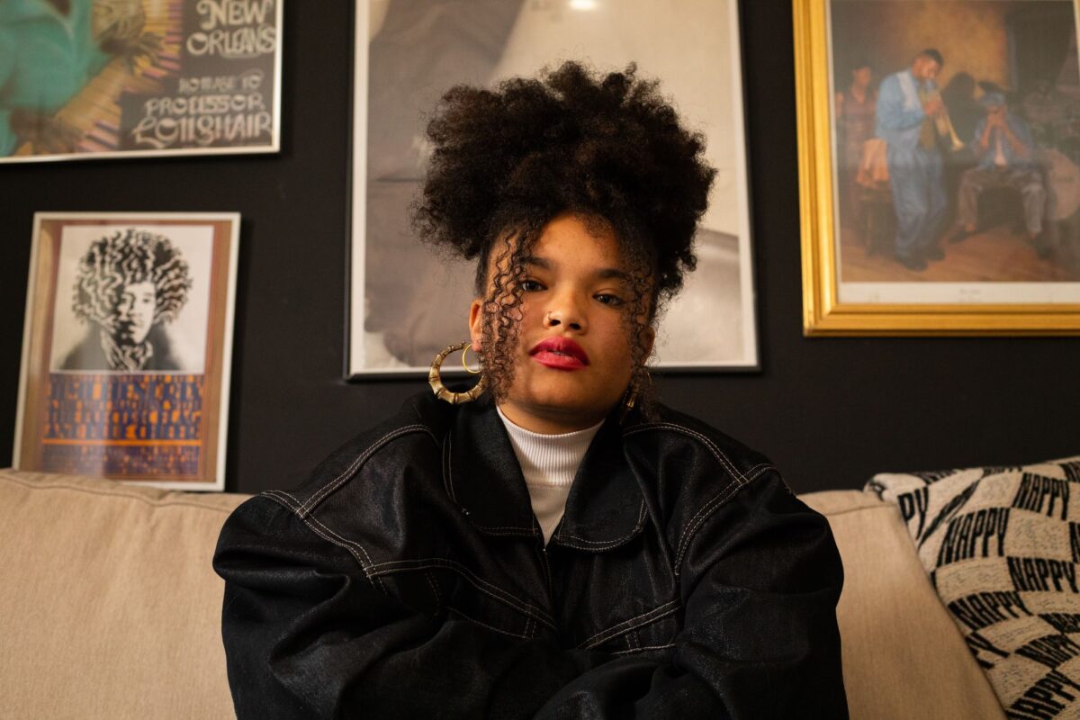 A person with curly hair and hoop earrings sits on a beige couch against a backdrop of framed artwork.