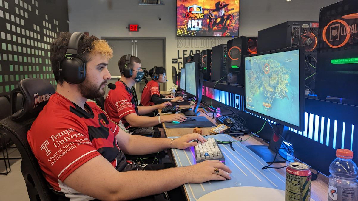 Gamers wearing headphones, seated at computer stations, focused on their screens. They are in a room with gaming posters on the wall and drinks on the table.