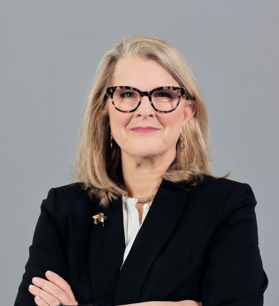 A woman with shoulder-length blonde hair, wearing glasses and a black blazer, stands with her arms crossed against a plain background.