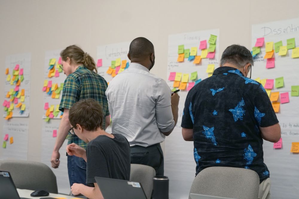Four people write on sticky notes attached to large sheets of paper on the wall in a meeting room. Two people standing, one sitting, and another partially visible are working collaboratively.