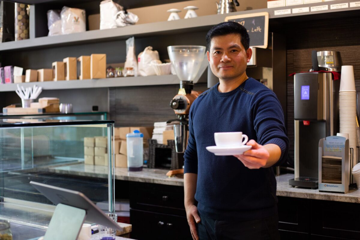 A person stands behind a coffee counter inside a café, holding out a white cup and saucer. The background shows various coffee-making equipment and supplies.