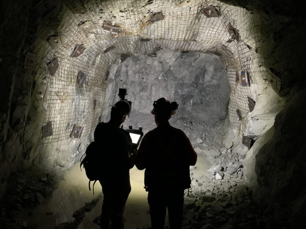 Two people wearing helmets stand in a dimly lit underground tunnel with rocky walls and a metal mesh overhead, using a tablet or device.