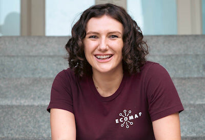A person with curly hair sits on steps, wearing a maroon shirt with the "ECOMAP" logo.