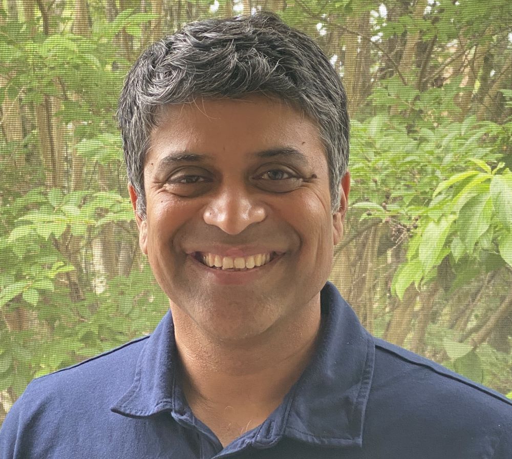 A person smiles at the camera. They are wearing a navy blue shirt with trees in the background.