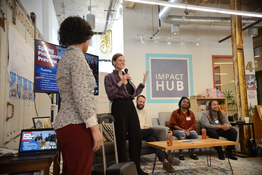 Michelle Geiss in purple shirt and black pants next to My-Azia Johnson in grey patterned shirt and red pants near people and wall with "Impact HUB" in white letters and purple square with blue outline
