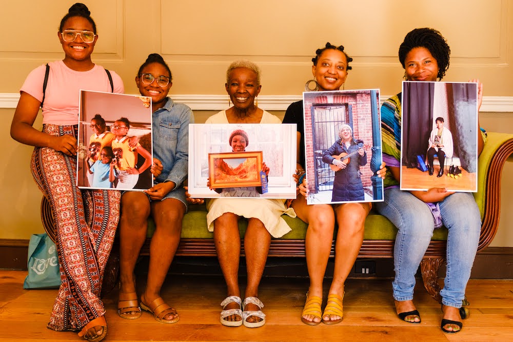 Women holding portraits while sitting on green bench before beige wall