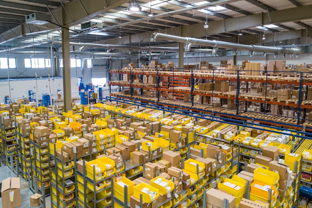 A large warehouse with numerous shelves filled with various boxes and packages, organized in rows under a high ceiling with industrial lighting.