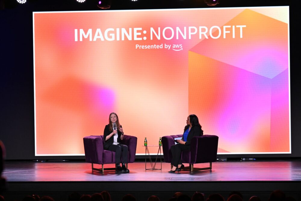 Chelsea Clinton sits in an armchair onstage across next to the interviewer, who also sits in an armchair. A large LED screen reading IMAGINE: NONPROFIT in white text on an orange background is behind them.