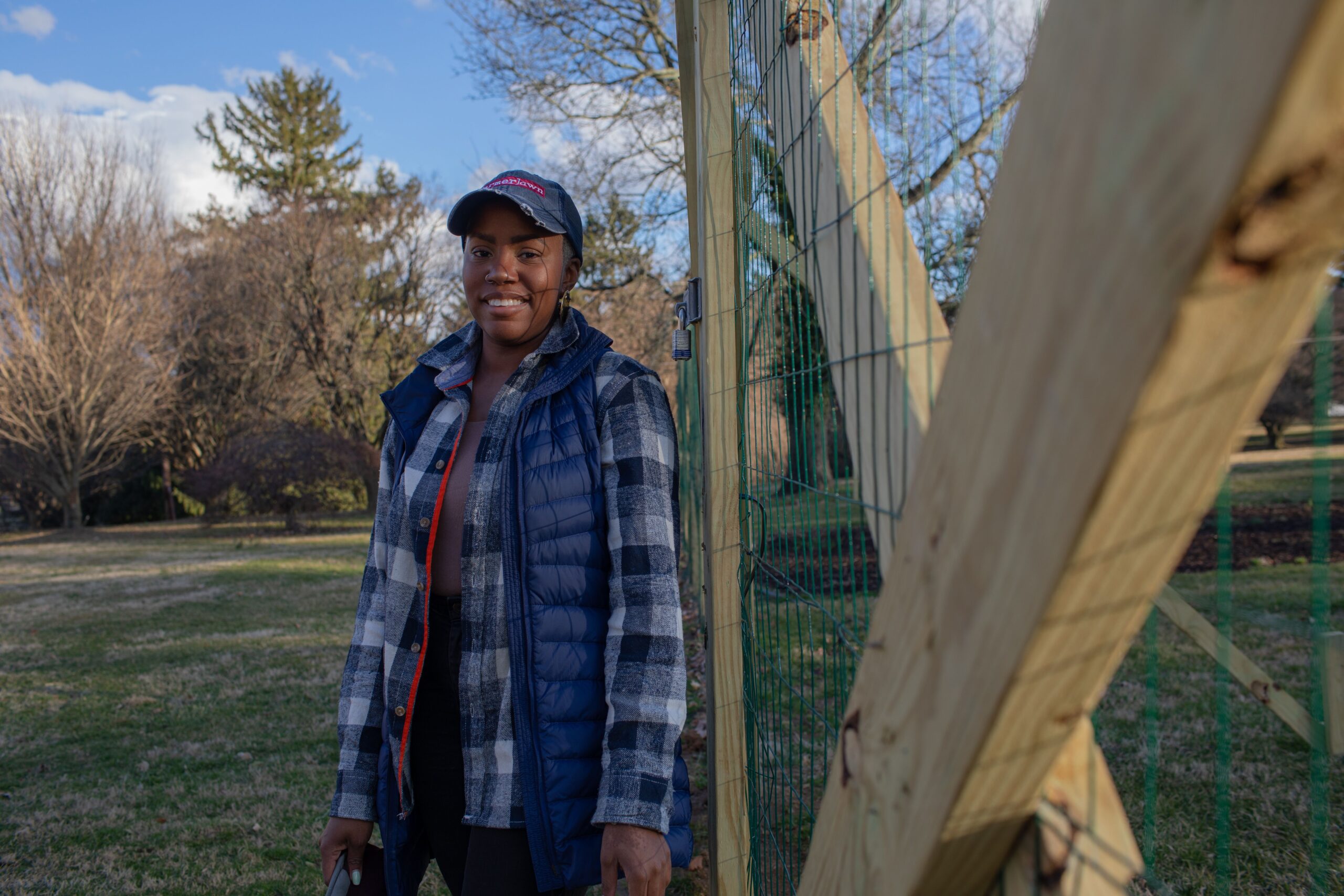 A person wearing a cap, checkered jacket, and blue vest stands outdoors near a wooden fence with trees and a grass field in the background.