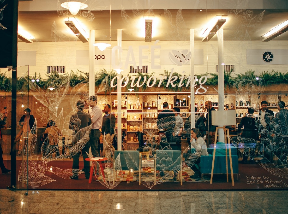 People working and socializing in a busy coworking space with bookshelves, plants, and cafe seating, seen through a glass front with "CAFÉ coworking" written on it.