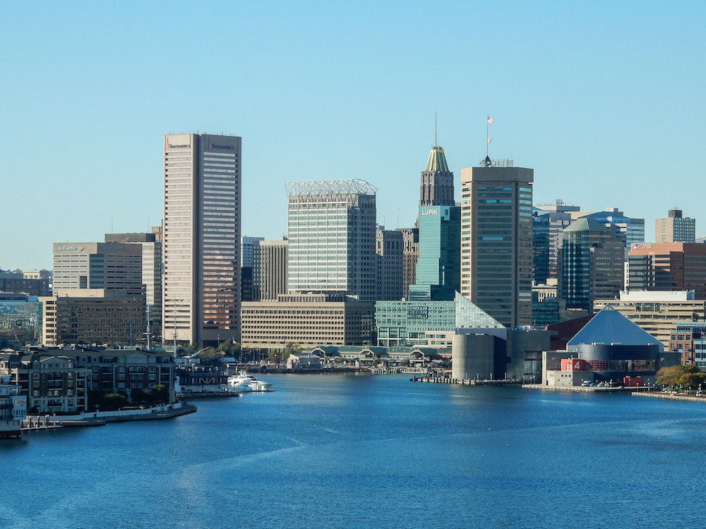 Multicolored buildings in Baltimore Skyline behind blue harbor water and before blue sky.