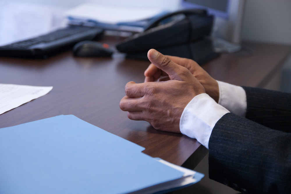 Hands of person in white shirt and charcoal pinstripe suit near blue folder on brown desk.
