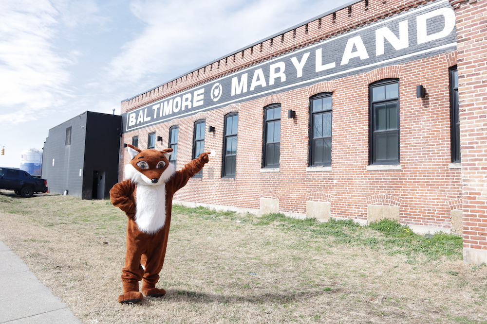 Person in brown fox outfit near red brick building that reads"BALTIMORE MARYLAND" in white text on black background near blue sky.