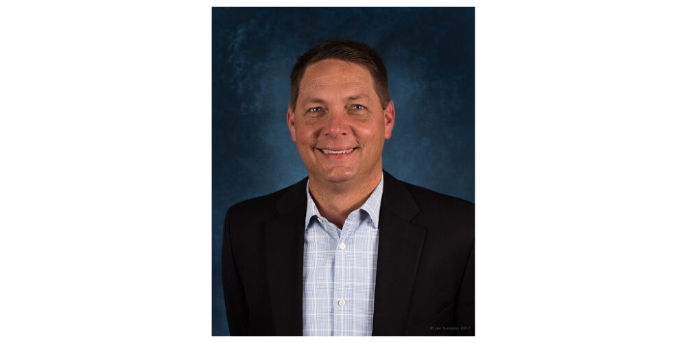 A smiling man wearing a dark jacket and shirt against a blue mottled background.
