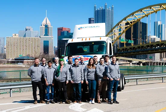 A group of people in matching gray jackets standing in front of a white truck with "optimus" written on it, with a city skyline and a yellow bridge in the background.