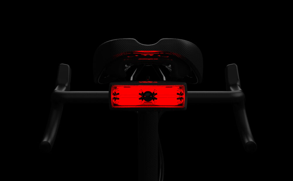 Red illuminated bike tail light with hazard symbol mounted under a black saddle, against a dark background.